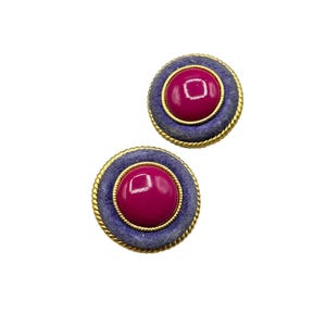 May include: A pair of round clip-on earrings. Each earring features a magenta center, surrounded by a blue velvet ring, and a gold rope-like trim. The earrings are set against a white background.
