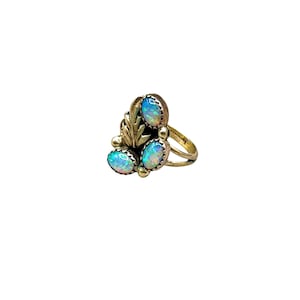 May include: A silver ring featuring three oval-shaped iridescent opals arranged in a cluster. The setting includes a decorative metal leaf motif positioned between the stones, with a split band design that tapers toward the back of the ring.