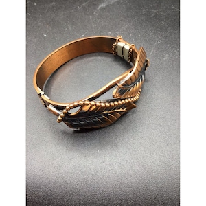 Puede incluir: Un brazalete de cobre con un diseño de hojas negras y doradas. El brazalete tiene un cierre de bisagra.