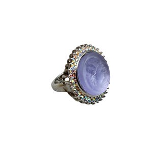 May include: Silver-toned ring featuring a light purple cameo with a profile portrait. The ring is surrounded by a halo of iridescent crystals. The ring's band has an ornate, openwork design.