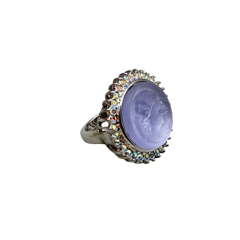 May include: Silver-toned ring featuring a light purple cameo with a profile portrait. The ring is surrounded by a halo of iridescent crystals. The ring's band has an ornate, openwork design.