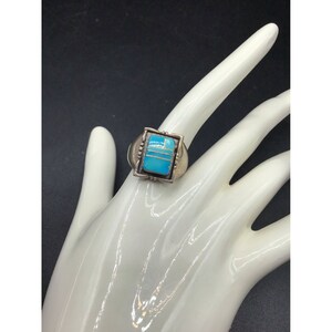 Puede incluir: Un anillo de plata con una piedra turquesa engastada en forma cuadrada. La piedra es de color azul claro y está rodeada por un borde de plata.