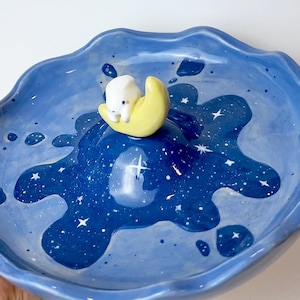 May include: A handmade ceramic dish with a wavy edge, painted in shades of blue to resemble a night sky. The dish features a central design of a white bear on a yellow crescent moon. White stars are scattered across the blue surface. The Etsy shop name is visible.