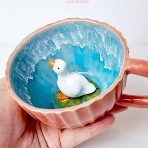 Op de afbeelding: Een keramische mok met een eendenfiguur erin. De mok heeft een koraalkleurige buitenkant en een blauwe binnenkant met een golfpatroon. De eend is wit met een oranje snavel, rustend op een groene waterlelie. De tekst "ceramiku" en "ceramikusshop.etsy.com" zijn zichtbaar.