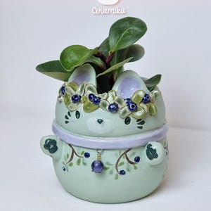 Puede incluir: Maceta de cerámica verde claro con forma de conejito, con una corona floral y un dije de cuentas moradas. Contiene una planta de hojas verdes. El texto "CeramikuShop.etsy.com" está en la parte inferior.