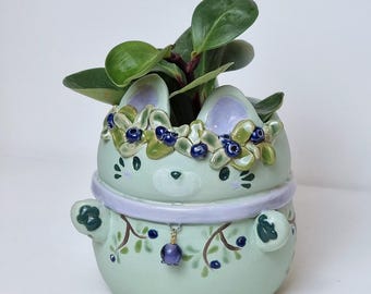 May include: A light green ceramic planter shaped like a bunny with a floral crown and a purple beaded charm. The planter is filled with a green leafy plant. The text "CeramikuShop.etsy.com" is at the bottom.