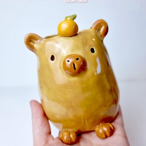 May include: A ceramic bear-shaped planter in a warm, golden-brown hue. The planter features a small, round orange fruit with a green stem on its head. The design includes small ears, a snout, and tiny feet. The text "Ceramiku" and "ceramikusshop.etsy.com" are visible.