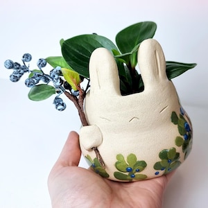 Könnte beinhalten: Ein cremefarbener Keramik-Pflanztopf in Hasenform, verziert mit grünen und blauen Blumenmustern, der einen Zweig mit künstlichen Beeren und Grünzeug hält. Der Topf ist mit grünen Blättern gefüllt.