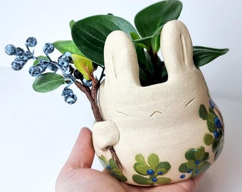May include: A cream-colored ceramic planter shaped like a bunny, holding a sprig of faux berries and greenery. The planter has green and blue floral accents. The bunny planter is filled with green leafy plants.