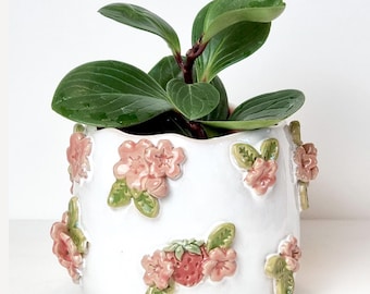 May include: A white ceramic planter with pink floral and green leaf embellishments, holding a green plant with large leaves. The planter has a glossy finish and the text "ceramiku shop etsy.com" is visible at the bottom.