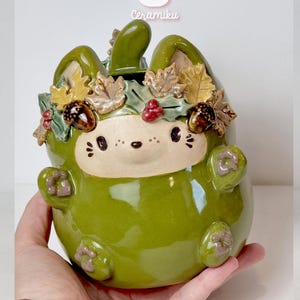 May include: A glossy green ceramic bunny-shaped container, decorated with a wreath of autumn leaves, acorns, and berries. The container has a white face with black details and the text "Ceramiku" and "ceramikusshop.etsy.com".