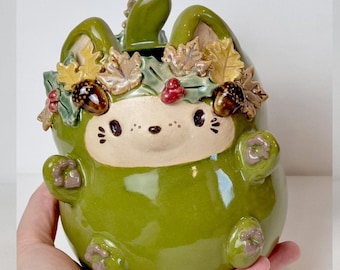 May include: A ceramic bunny-shaped container in glossy green, adorned with a fall-themed wreath of leaves, acorns, and berries. The container has a white face with black details and the text "Ceramiku" and "ceramikusshop.etsy.com".