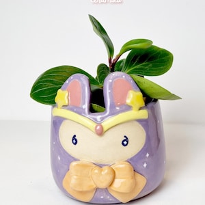 Könnte beinhalten: Ein lavendelfarbener Keramik-Pflanztopf in Hasenform mit gelber Schleife und Sternen. Er enthält eine kleine grüne Pflanze mit großen Blättern. Der Text "Ceramiku" und "ceramikusshop.etsy.com" sind sichtbar.