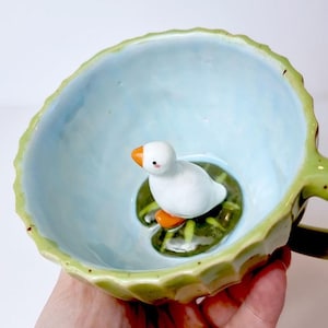 Pode incluir: Uma chávena de cerâmica com interior azul claro e exterior verde em forma de folha. No interior, um pato de cerâmica branco com patas e bico laranja repousa sobre uma folha de nenúfar verde. A chávena é segurada por uma mão.