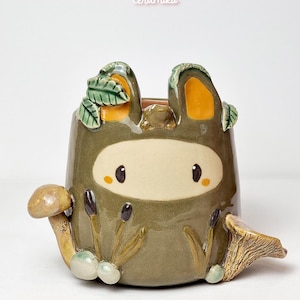 May include: A handmade ceramic planter with a whimsical design. The planter is olive green with a cartoon-like face and mushroom accents. The text "Ceramiku" and "ceramikusshop.etsy.com" are visible.