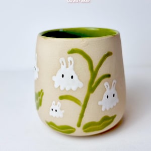 May include: A beige ceramic mug with a green interior and a design of white bunny figures and green plant stems. The mug has the text "Ceramiku" and "ceramikusshop.etsy.com".