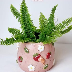 Può includere: Un vaso di ceramica rosa con una felce. Il vaso è decorato con fiori bianchi, fragole rosse, foglie verdi e una piccola statuina di gatto. Il testo "Ceramiku" e "ceramikusshop.etsy.com" sono visibili.