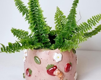 May include: A pink ceramic planter with a fern plant. The planter is decorated with white flowers, red strawberries, green leaves, and a small cat figurine. The text "Ceramiku" and "ceramikusshop.etsy.com" are visible.
