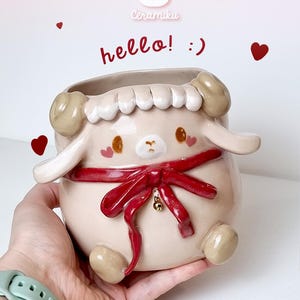 May include: A sheep-shaped ceramic planter in beige, with white wool detailing and a red ribbon. The planter features a cute face with brown eyes and the text "hello! :)" is written on the side.
