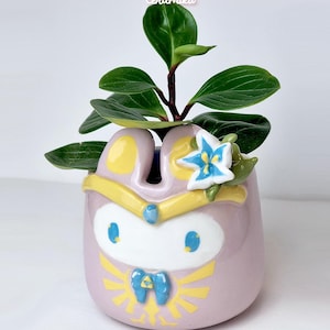 Puede incluir: Una maceta de cerámica de color lila claro, con un diseño de personaje de dibujos animados con detalles amarillos y azules. Contiene una pequeña planta verde con hojas anchas. El texto "Ceramiku" y "ceramikusshop.etsy.com" son visibles.