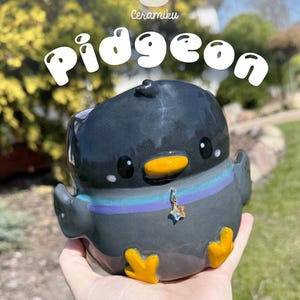 May include: A ceramic pigeon-shaped mug with the word "Pidgeon" in white script. The mug is primarily dark gray with yellow feet and beak, and a blue, purple, and teal striped collar. A small star charm hangs from the collar.