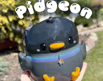 Op de afbeelding: Een keramische mok in de vorm van een duif met het woord "Pidgeon" in witte letters. De mok is voornamelijk donkergrijs met gele voeten en snavel, en een blauwe, paarse en blauwgroene gestreepte kraag. Een kleine sterrenhanger hangt aan de kraag.