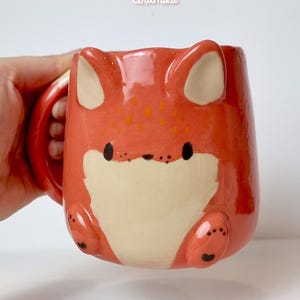 K&ouml;nnte beinhalten: Keramikbecher in Fuchsform mit orangefarbenen und cremefarbenen Farben. Der Becher hat einen Henkel und das Gesicht des Fuchses ist auf der Vorderseite. Der Text "Ceramiku" steht oben und "ceramikushop.etsy.com" unten.