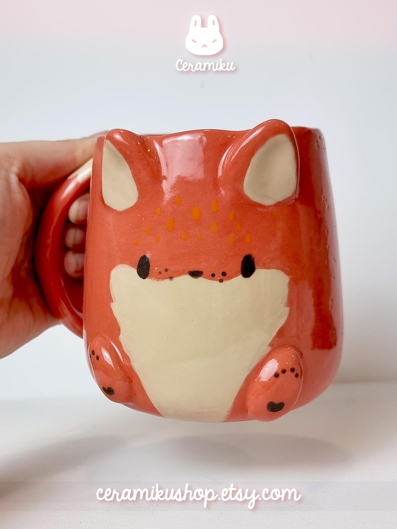 K&ouml;nnte beinhalten: Keramikbecher in Fuchsform mit orangefarbenen und cremefarbenen Farben. Der Becher hat einen Henkel und das Gesicht des Fuchses ist auf der Vorderseite. Der Text "Ceramiku" steht oben und "ceramikushop.etsy.com" unten.