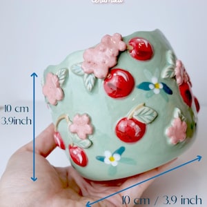 Pode incluir: Um vaso de cer&acirc;mica verde claro, com 10 cm de largura, adornado com cerejas vermelhas em relevo e flores rosa. O vaso tem uma borda ondulada e &eacute; decorado com detalhes florais brancos e azuis. O texto "Ceramiku" e "ceramikusshop.etsy.com" s&atilde;o vis&iacute;veis.