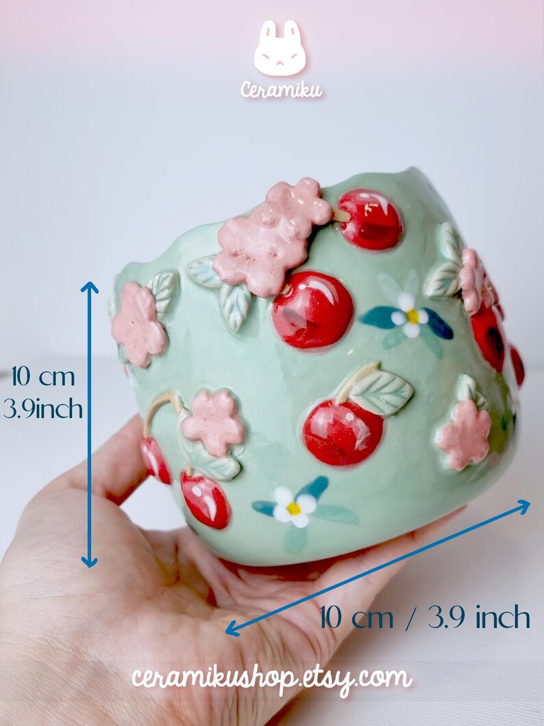 Pode incluir: Um vaso de cer&acirc;mica verde claro, com 10 cm de largura, adornado com cerejas vermelhas em relevo e flores rosa. O vaso tem uma borda ondulada e &eacute; decorado com detalhes florais brancos e azuis. O texto "Ceramiku" e "ceramikusshop.etsy.com" s&atilde;o vis&iacute;veis.