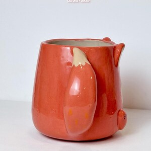 K&ouml;nnte beinhalten: Eine Keramik-Tasse in leuchtendem Orange, geformt wie ein Fuchs. Die Tasse hat einen Fuchsschwanz als Griff und kleine Ohrendetails. Der Text "Ceramiku" und "ceramikushop.etsy.com" sind sichtbar.