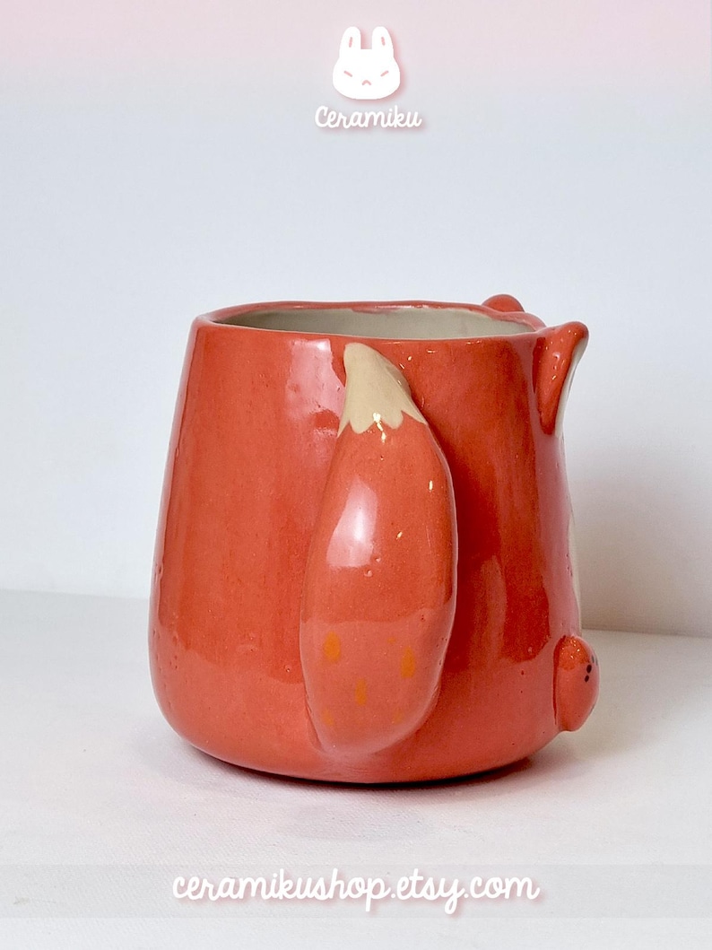K&ouml;nnte beinhalten: Eine Keramik-Tasse in leuchtendem Orange, geformt wie ein Fuchs. Die Tasse hat einen Fuchsschwanz als Griff und kleine Ohrendetails. Der Text "Ceramiku" und "ceramikushop.etsy.com" sind sichtbar.