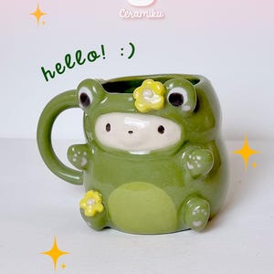 Könnte beinhalten: Grüner Keramikbecher in Froschform mit weißem Gesicht und gelben Blumenakzenten. Der Becher hat einen Henkel und den Text "hello! :)". Der Markenname "Ceramiku" ist ebenfalls sichtbar. Ein niedliches und skurriles Design.