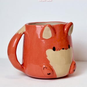 K&ouml;nnte beinhalten: Keramikbecher in Fuchsform, orange und cremefarben. Der Becher hat ein Fuchgesicht, Ohren und einen Schwanz. Die Texte "Ceramiku" und "ceramikusshop.etsy.com" sind sichtbar. Ein charmantes und einzigartiges Design.