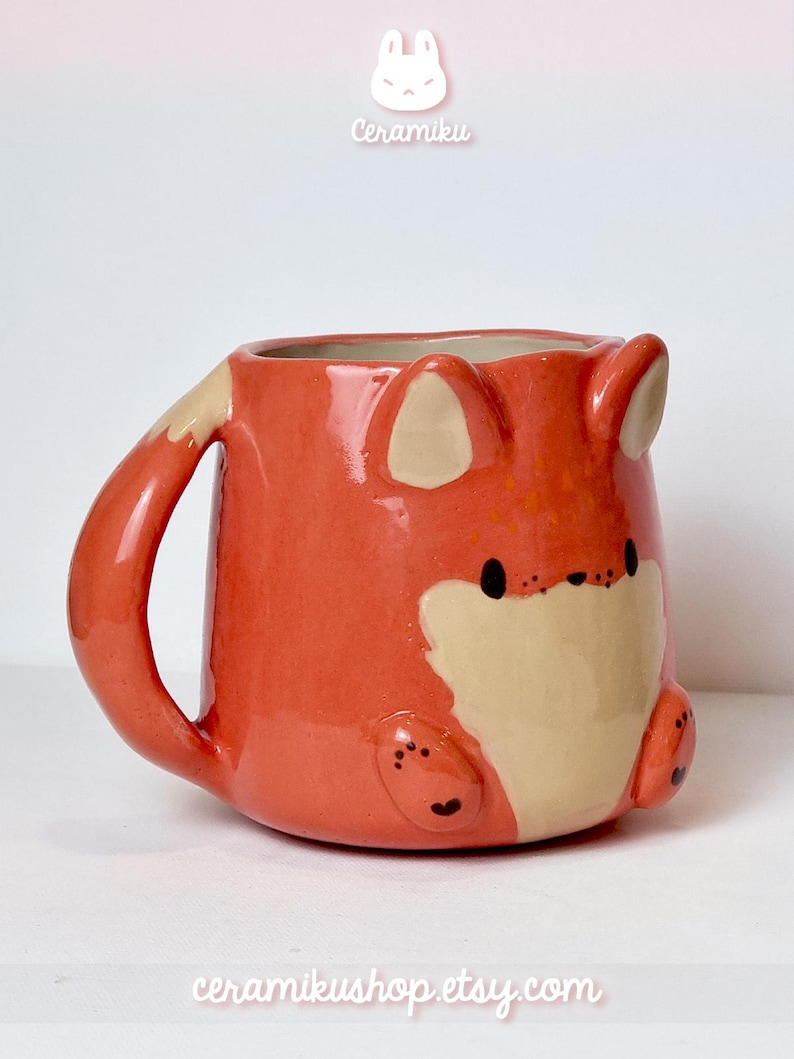 K&ouml;nnte beinhalten: Keramikbecher in Fuchsform, orange und cremefarben. Der Becher hat ein Fuchgesicht, Ohren und einen Schwanz. Die Texte "Ceramiku" und "ceramikusshop.etsy.com" sind sichtbar. Ein charmantes und einzigartiges Design.