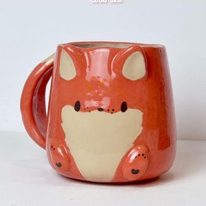 K&ouml;nnte beinhalten: Eine keramische Tasse in Form eines Fuchses, in Orange und Creme. Die Tasse hat Fuchseigenschaften, einschlie&szlig;lich Ohren, Pfoten und einem Schwanz. Die Texte "Ceramiku" und "ceramikusshop.etsy.com" sind sichtbar.