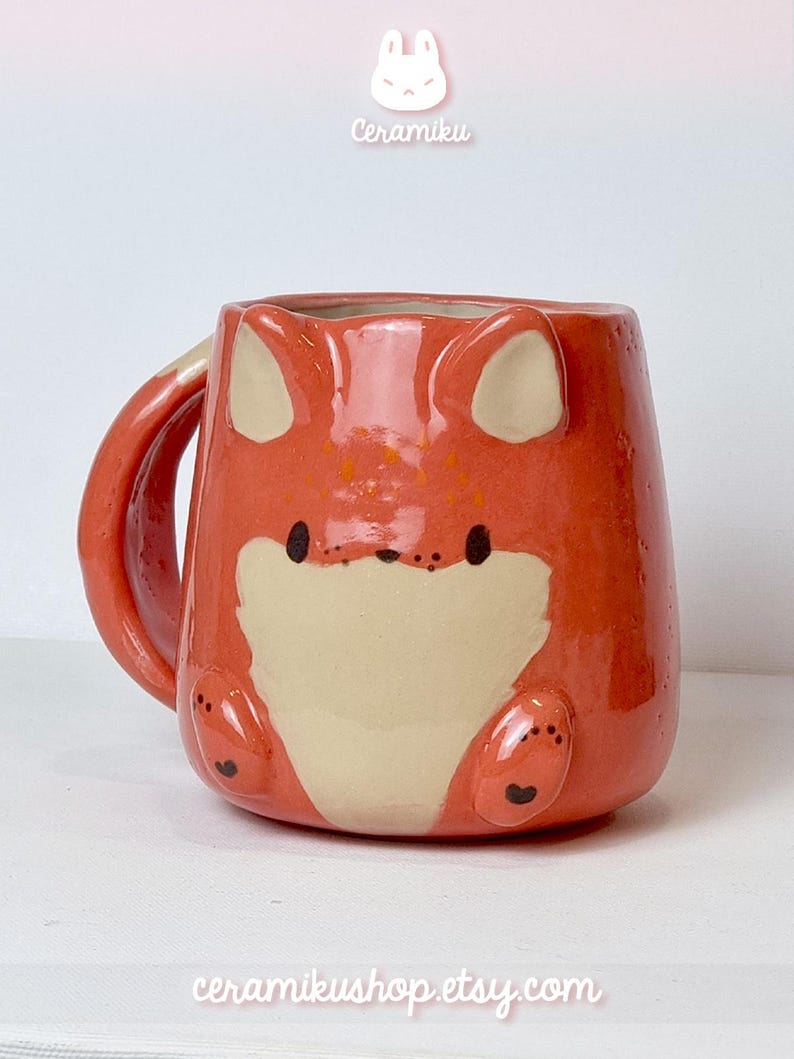 K&ouml;nnte beinhalten: Eine keramische Tasse in Form eines Fuchses, in Orange und Creme. Die Tasse hat Fuchseigenschaften, einschlie&szlig;lich Ohren, Pfoten und einem Schwanz. Die Texte "Ceramiku" und "ceramikusshop.etsy.com" sind sichtbar.