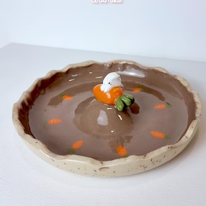 Può includere: Un piatto in ceramica con interno marrone e bordo beige maculato. Il piatto presenta un cumulo centrale con una carota e una piccola statuina di coniglietto bianco. Piccoli disegni di carote arancioni sono dipinti all'interno. I testi "Ceramiku" e "ceramikusshop.etsy.com" sono visibili.