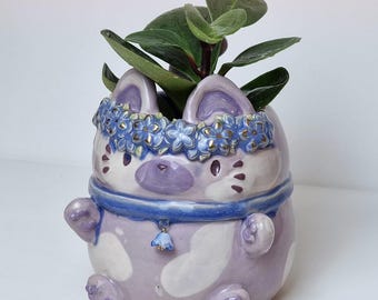 May include: A whimsical ceramic planter in the shape of a cat, painted in lavender and white with blue floral accents. The planter features a blue collar with a bell charm and is filled with green foliage. The text "CeramikuShop.etsy.com" is visible.