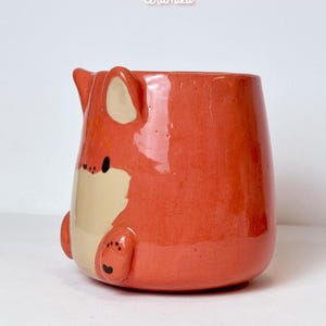 K&ouml;nnte beinhalten: Keramikbecher in Fuchsform, in Orange und Creme. Der Becher hat ein Fuchskopf-Design mit Ohren, Nase und Pfoten. Die Texte "Ceramiku" und "ceramikusshop.etsy.com" sind sichtbar.