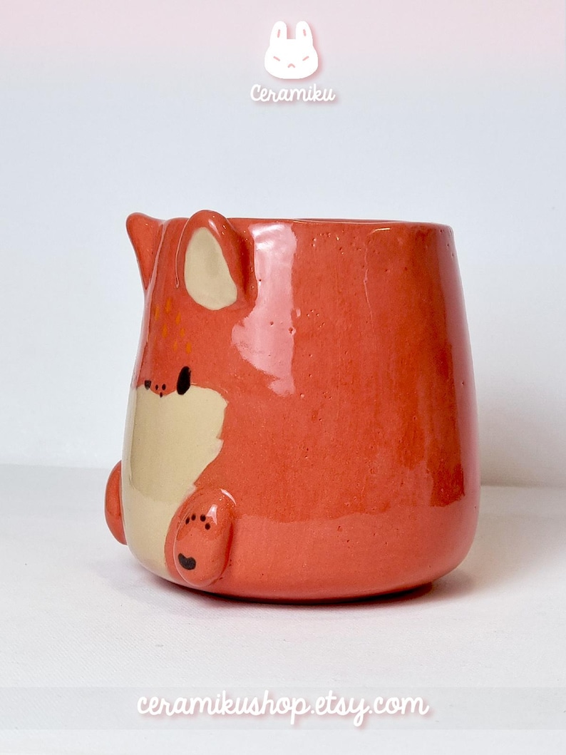 K&ouml;nnte beinhalten: Keramikbecher in Fuchsform, in Orange und Creme. Der Becher hat ein Fuchskopf-Design mit Ohren, Nase und Pfoten. Die Texte "Ceramiku" und "ceramikusshop.etsy.com" sind sichtbar.
