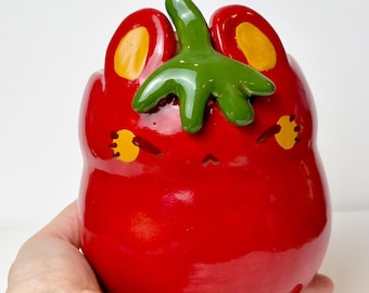 May include: A red ceramic tomato-shaped container with a green stem and yellow accents. The container has a cute face design and the text "Ceramiku" and "ceramikusshop.etsy.com".