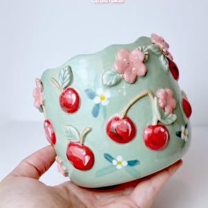 Puede incluir: Un cuenco de cerámica verde menta decorado con cerezas rojas, flores rosas y flores blancas. El cuenco tiene un borde ondulado y se sostiene en una mano. El nombre de la tienda "Ceramiku" y la URL de Etsy de la tienda son visibles.