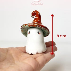 Puede incluir: Una figura de hongo de cerámica, de aproximadamente 8 cm de altura, con un cuerpo blanco y un sombrero marrón rojizo salpicado de puntos blancos. El sombrero tiene una forma curvada y una parte inferior verde. La figura tiene pequeñas mejillas rosadas. La figura se sostiene en una mano.