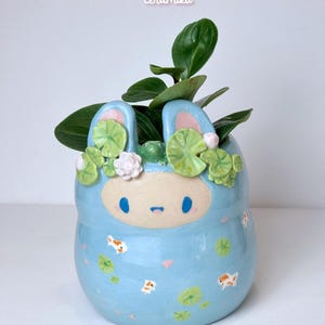 May include: A light blue ceramic planter shaped like a bunny with a smiling face, ears, and floral accents. The planter features koi fish and lily pads, and holds green plants. The text "Ceramiku" and "ceramikushop.etsy.com" are visible.