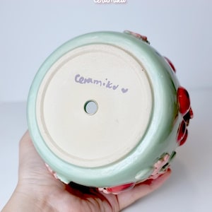 Pode incluir: Jarro de cer&acirc;mica verde claro com interior branco. O jarro apresenta detalhes florais vermelhos e rosa. O texto "Ceramiku" est&aacute; escrito na base. O URL da loja Etsy tamb&eacute;m &eacute; vis&iacute;vel.