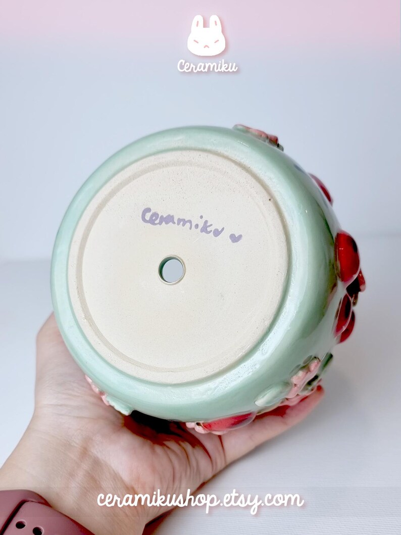 Pode incluir: Jarro de cer&acirc;mica verde claro com interior branco. O jarro apresenta detalhes florais vermelhos e rosa. O texto "Ceramiku" est&aacute; escrito na base. O URL da loja Etsy tamb&eacute;m &eacute; vis&iacute;vel.