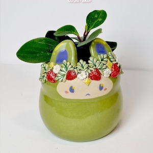 Könnte beinhalten: Ein grüner Keramik-Pflanztopf mit einem Cartoon-Gesicht, blauen Akzenten und einem Erdbeer- und Blumenkranz. Der Topf enthält eine kleine grüne Pflanze mit großen Blättern. Der Etsy-Shop-Name "ceramiku" steht oben und "ceramukishop.etsy.com" unten.