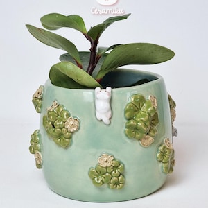 Op de afbeelding: Een lichtgroene keramische plantenpot met een klein wit konijnfiguur en reliëf bloemmotieven. De pot bevat een groene bladplant. De teksten "Ceramiku" en "CeramikuShop.etsy.com" zijn zichtbaar.