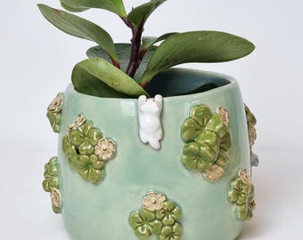 Op de afbeelding: Een lichtgroene keramische plantenpot met een klein wit konijnfiguur en reliëf bloemmotieven. De pot bevat een groene bladplant. De teksten "Ceramiku" en "CeramikuShop.etsy.com" zijn zichtbaar.